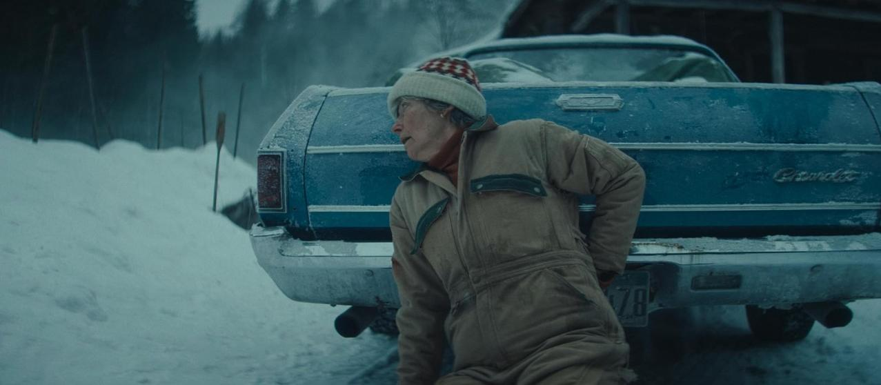 Eine Person in einem braunen Overall und Mütze steht neben einem blauen Auto im Schnee.