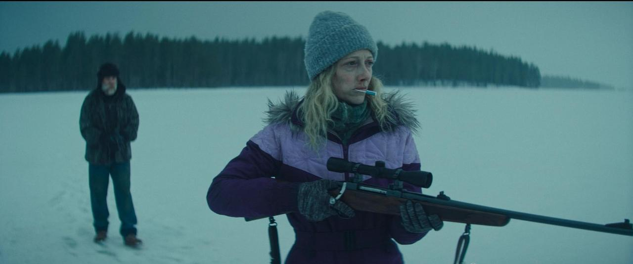 Eine Frau mit einem Lolli hält ein Gewehr in einer verschneiten Landschaft. Ein Mann steht im Hintergrund.