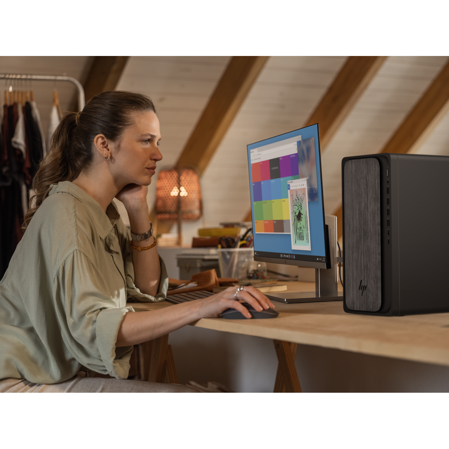 Woman at desk using computer in a room with slanted ceiling. Clothes rack in background.