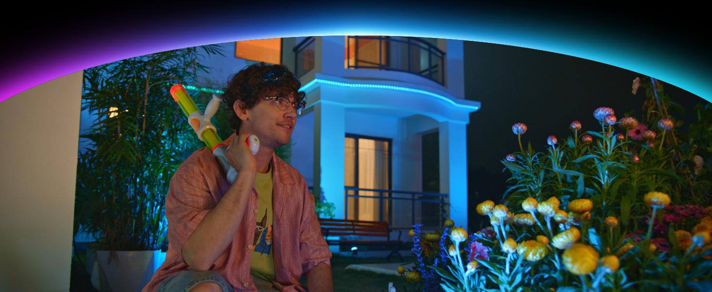 Ein Mann mit Brille und Locken hält eine Wasserpistole in einem Garten mit beleuchtetem Haus.