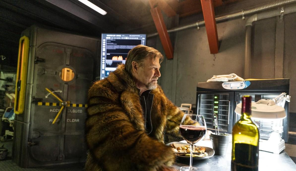 Ein Mann in einem Pelzmantel sitzt an einem Tisch mit Wein und Essen. Eine große Tür ist hinter ihm.