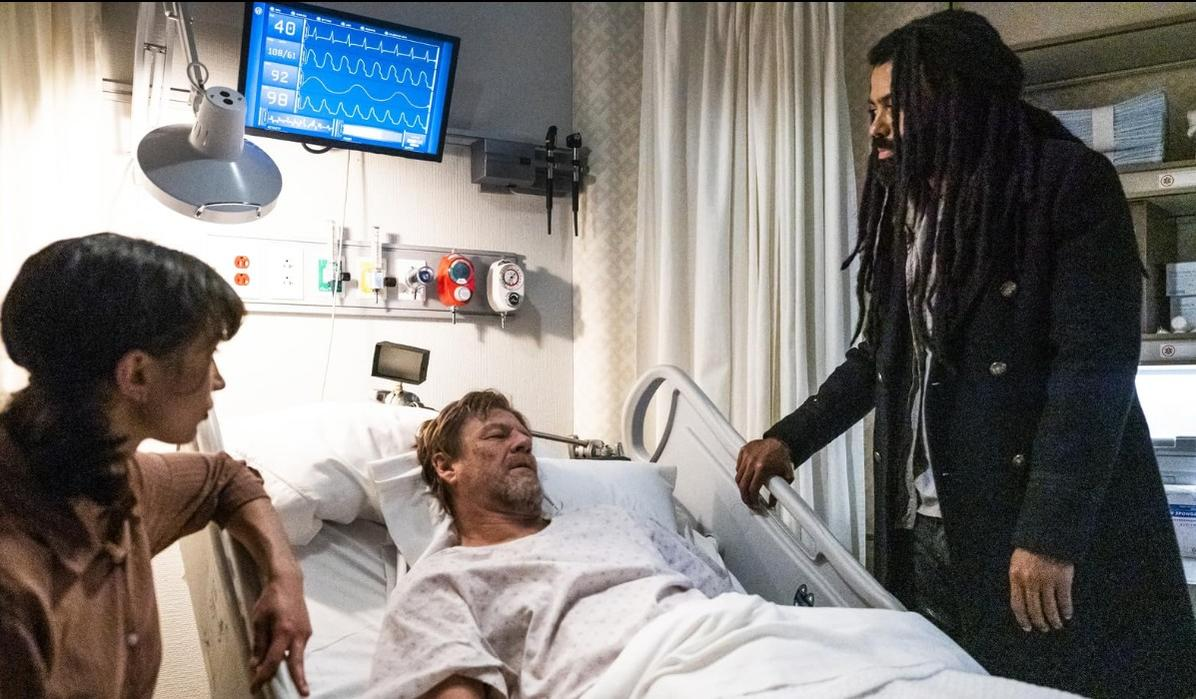 Ein Mann im Krankenhausbett, beobachtet von einer Frau und einem Mann mit Dreadlocks.