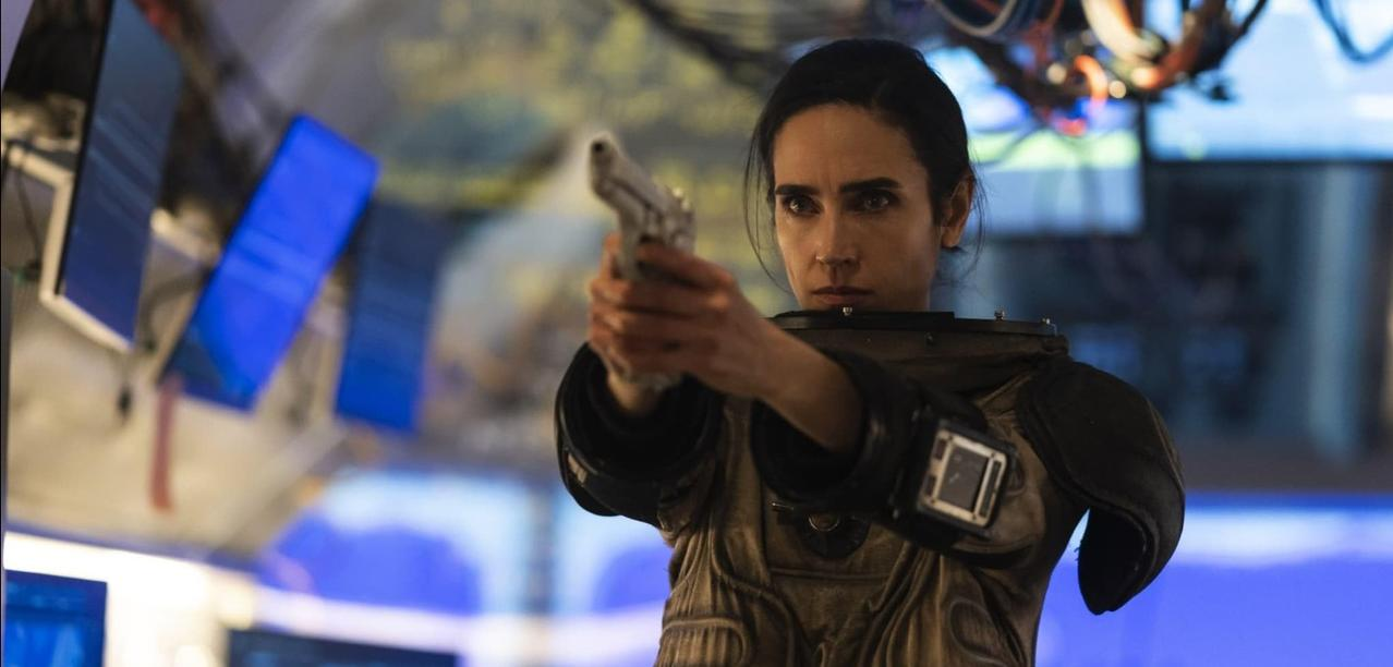 Eine Frau in einem futuristischen Anzug hält eine Waffe und zielt nach vorne in einem schwach beleuchteten Raum mit blauen Bildschirmen.