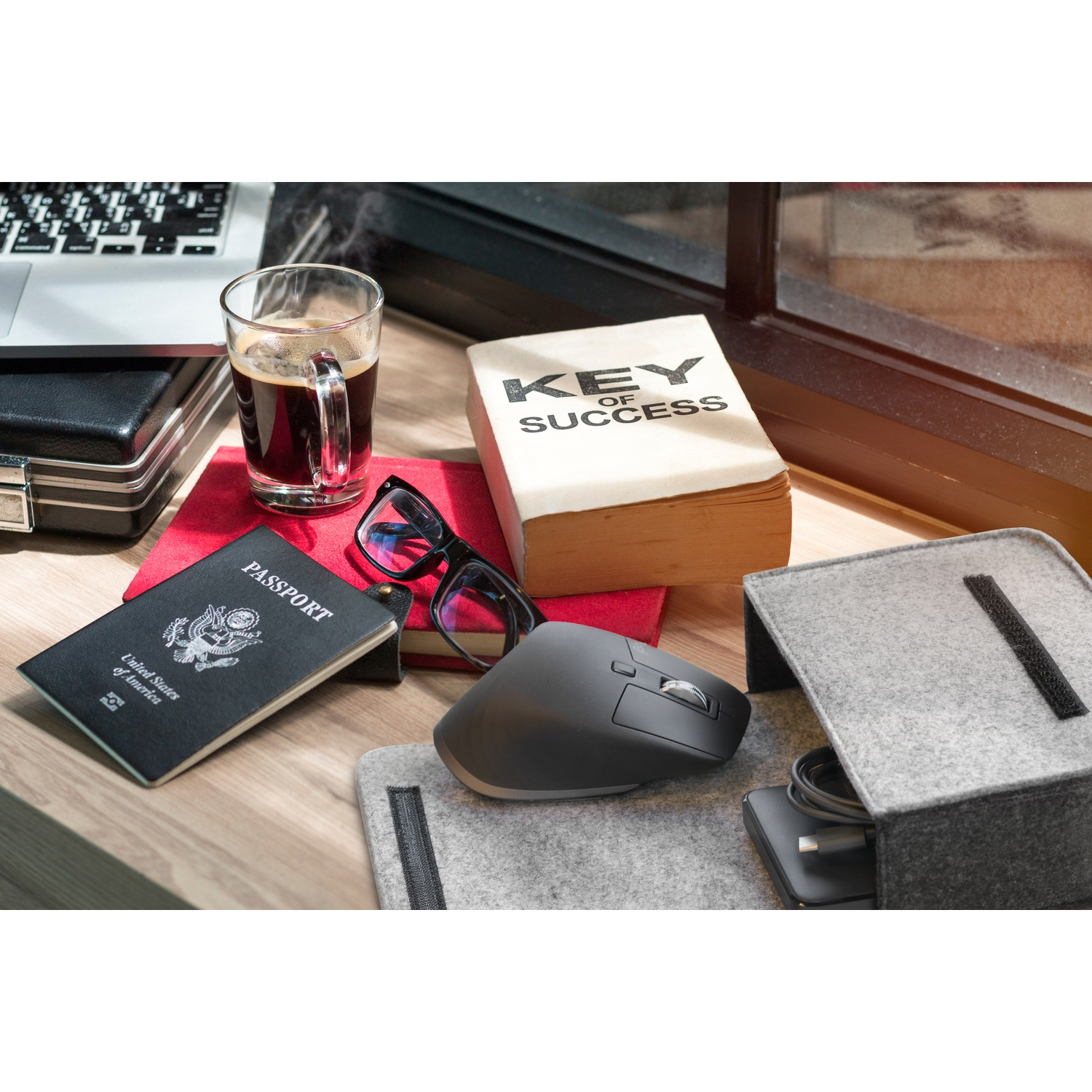 Scrivania con laptop, caffè, passaporto, occhiali, libro, mouse e contenitore.