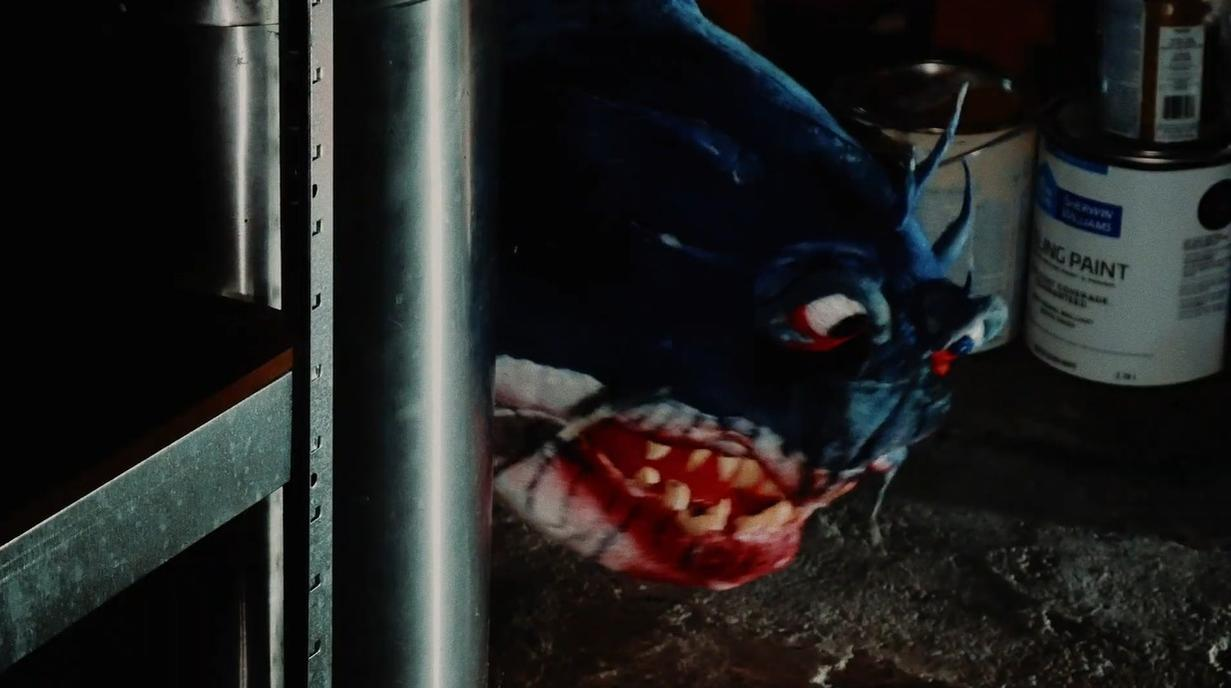 Ein blaues Monster mit roten Augen und spitzen Zähnen sitzt neben Farbeimern.