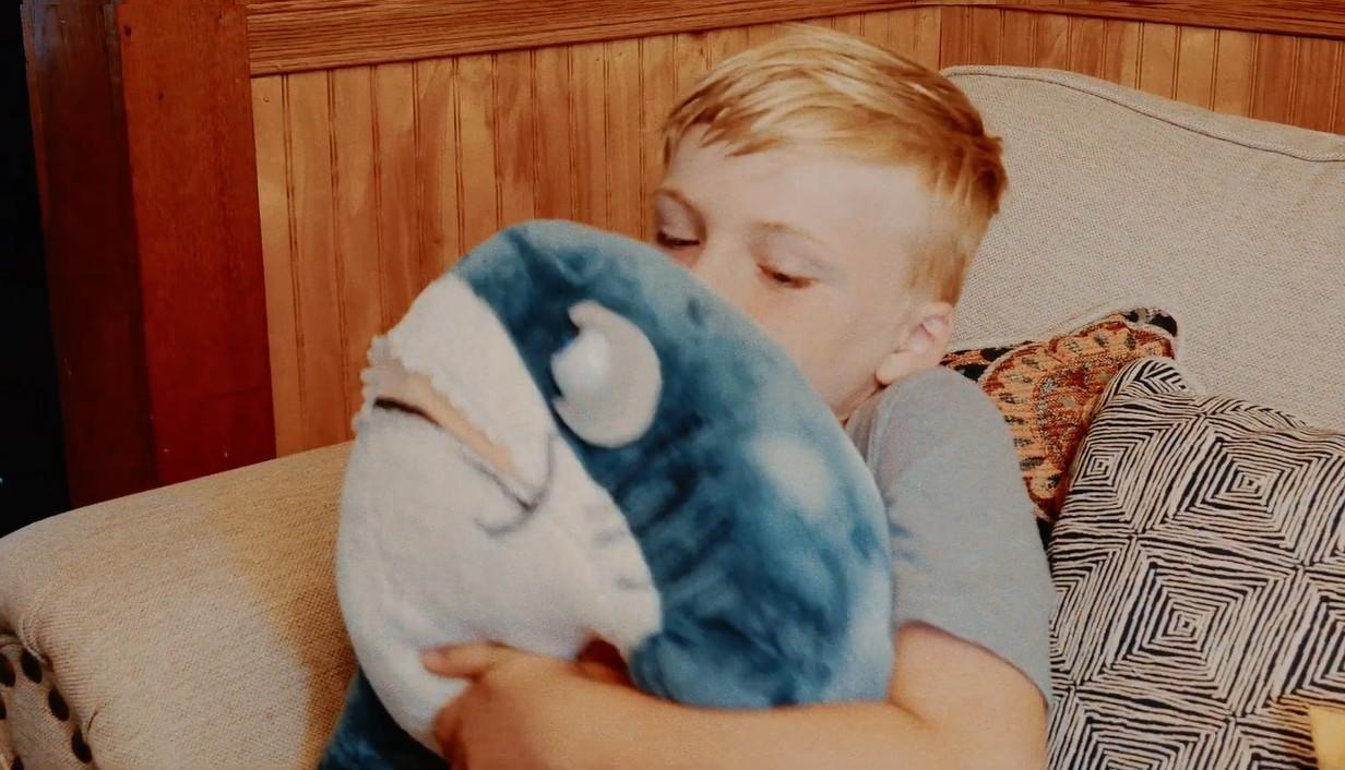 Ein kleiner Junge umarmt einen blau-weißen Plüschhai und sitzt auf einer Couch mit gemusterten Kissen.