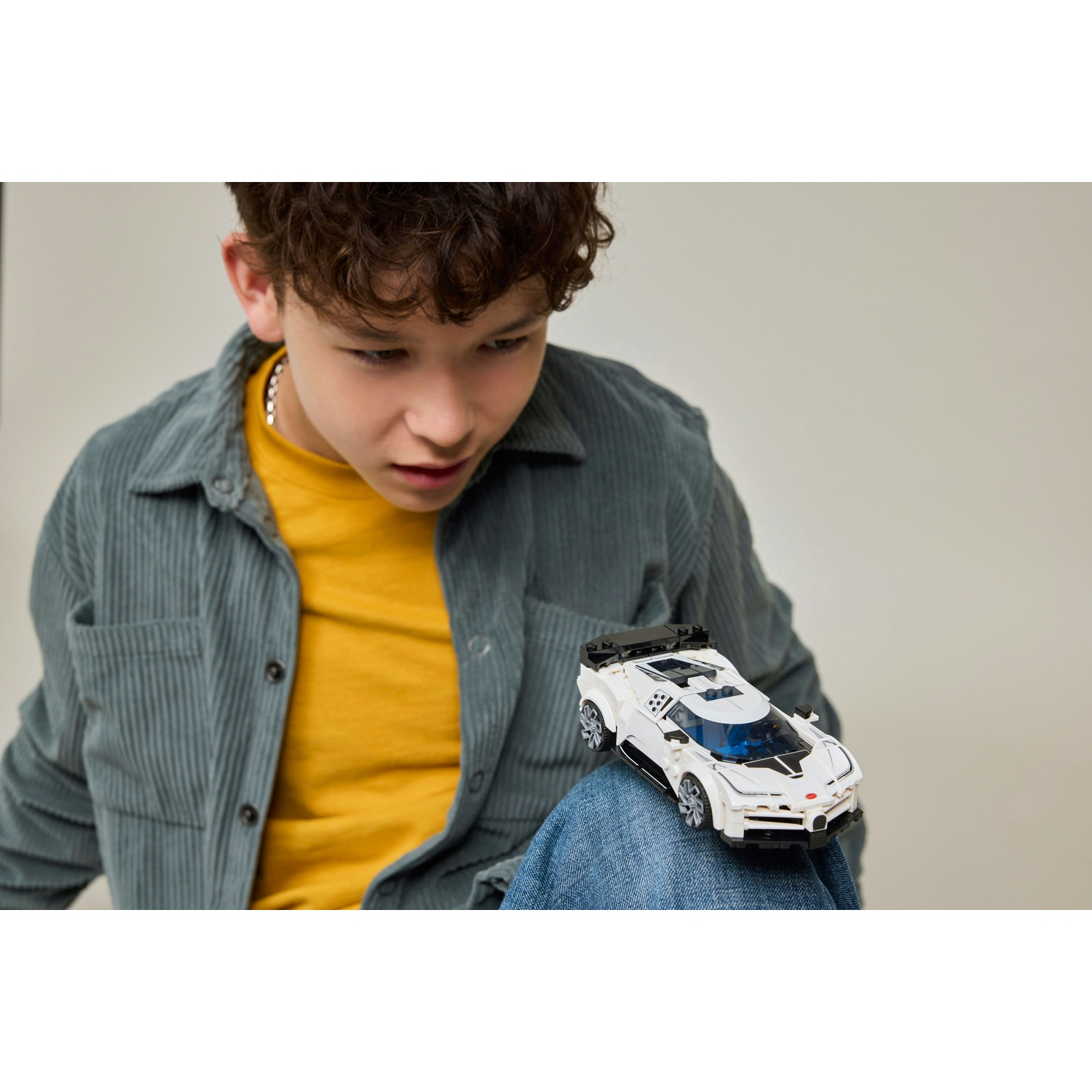 Un ragazzo con giacca di velluto a coste grigia e maglietta gialla guarda un'auto giocattolo bianca sui suoi jeans blu.