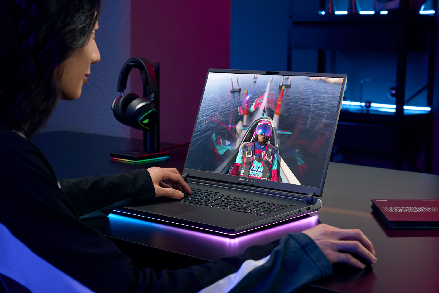 Person plays racing game on laptop. Headphones on stand nearby. RGB lighting.