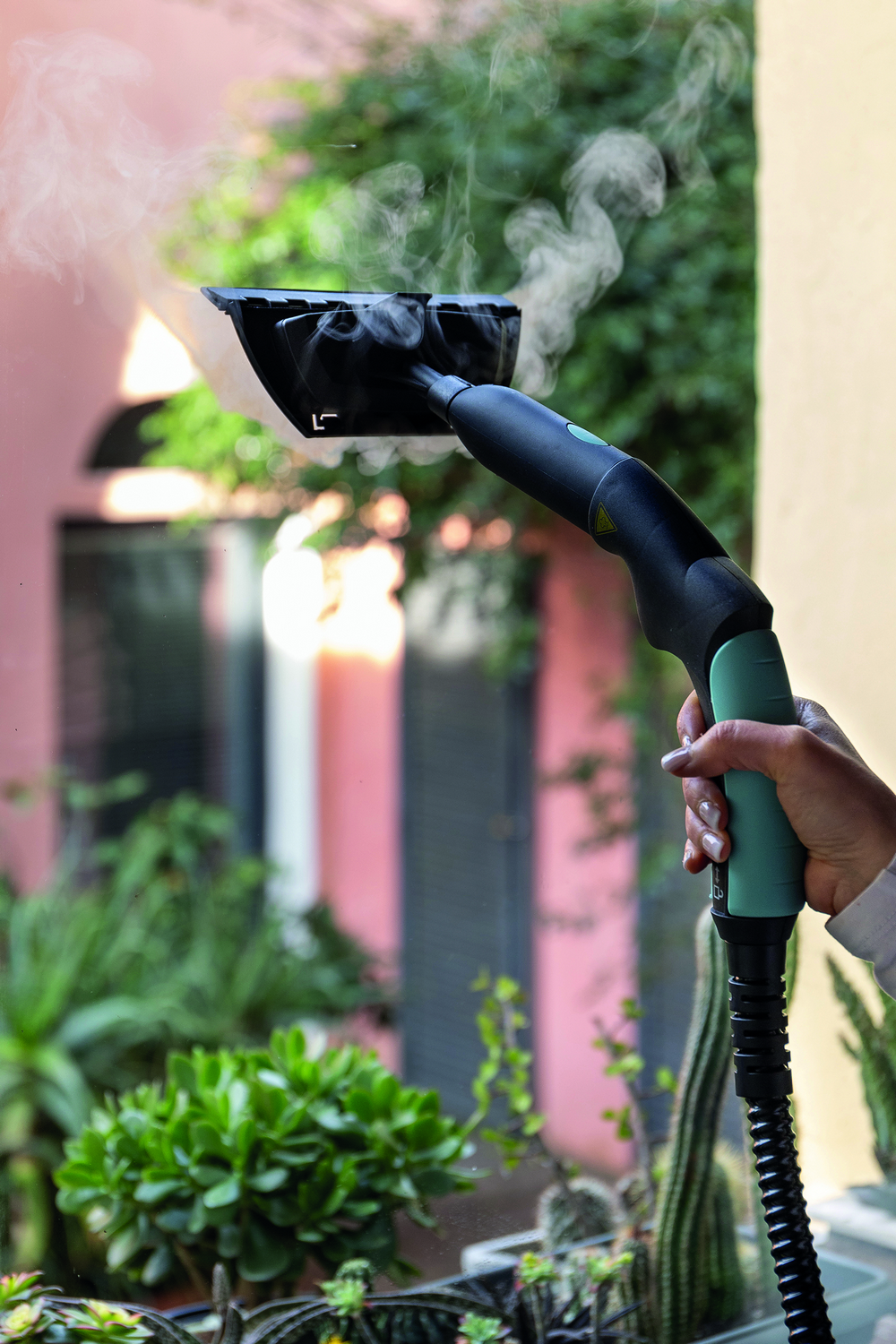 Una mano sostiene una boquilla de limpiador a vapor, rociando vapor. Se ven plantas y un edificio rosa.