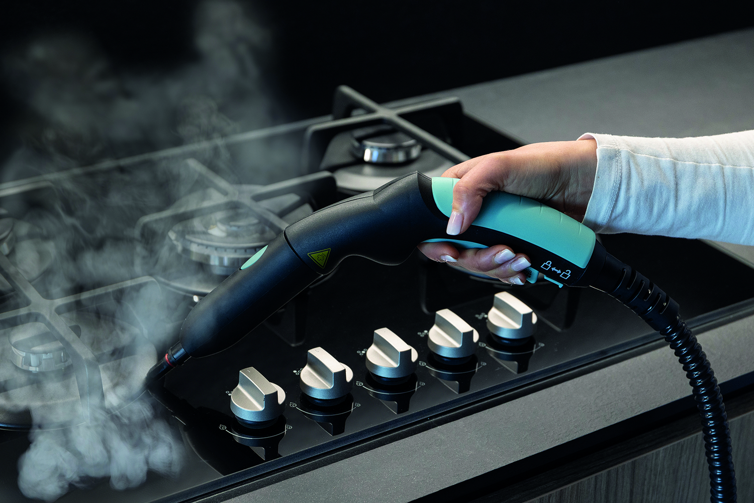 Mano sujetando boquilla limpiadora a vapor rociando vapor sobre cocina de gas.
