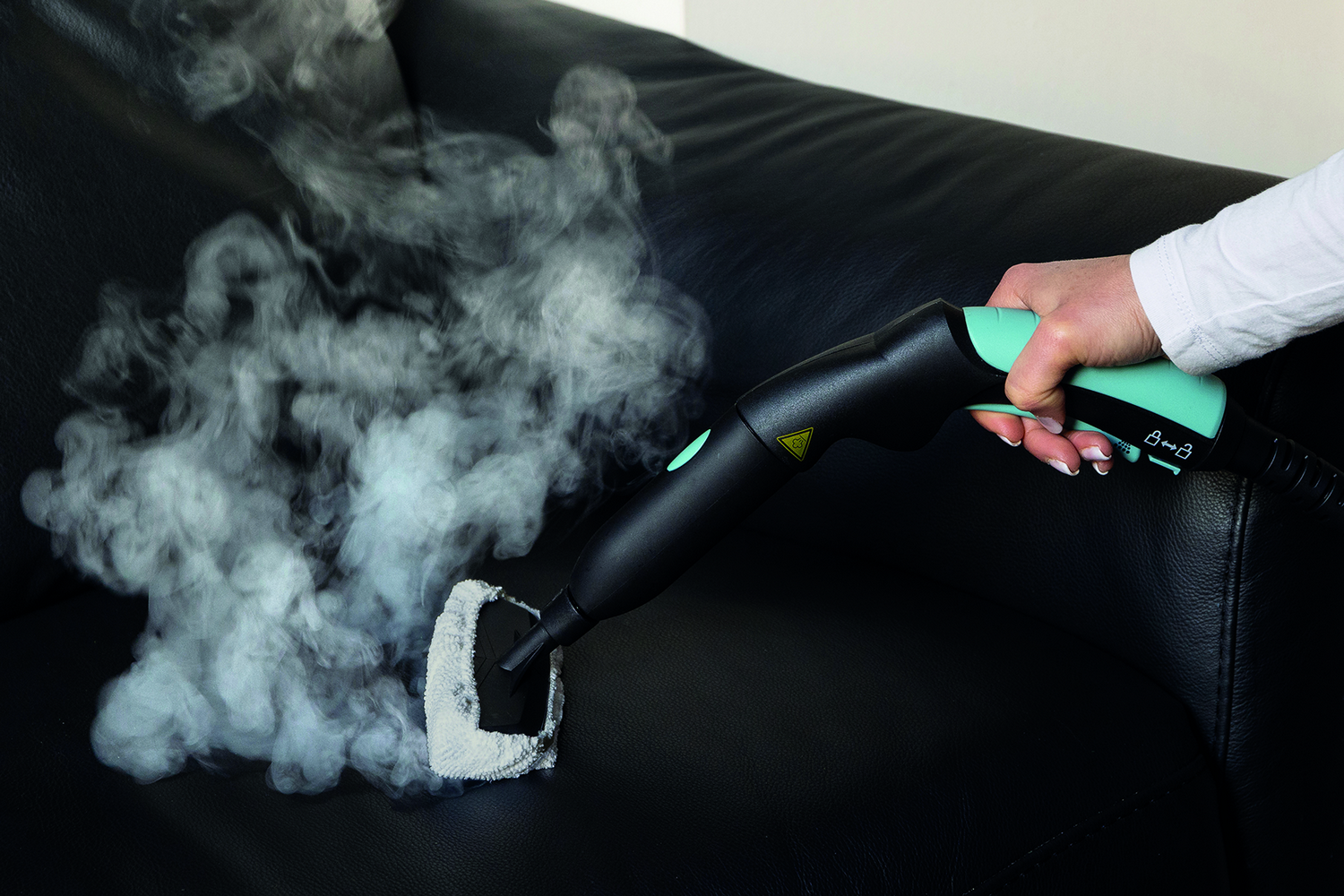 Una mano sostiene un limpiador a vapor negro y turquesa, vaporizando un sofá de cuero negro.