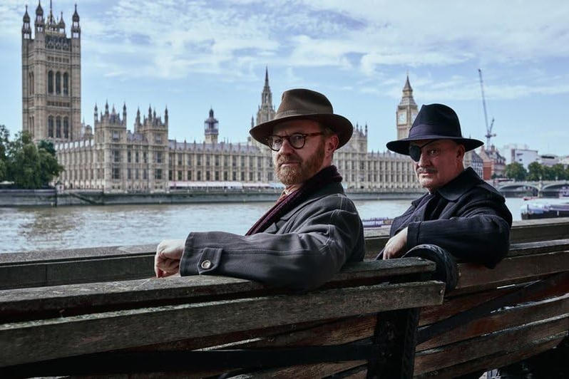 Zwei Männer mit Hüten sitzen auf einer Bank vor den Houses of Parliament in London.