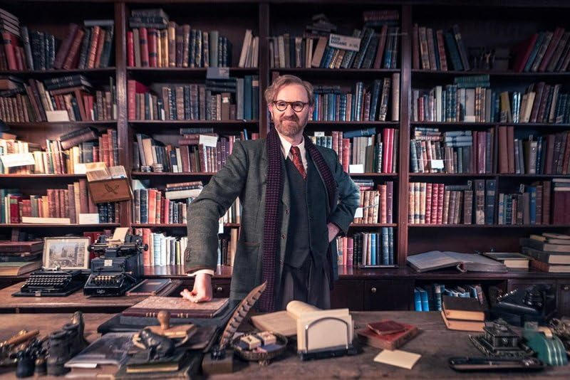 Mann mit Brille und Anzug steht am Schreibtisch mit Schreibmaschinen und Bücherregalen.