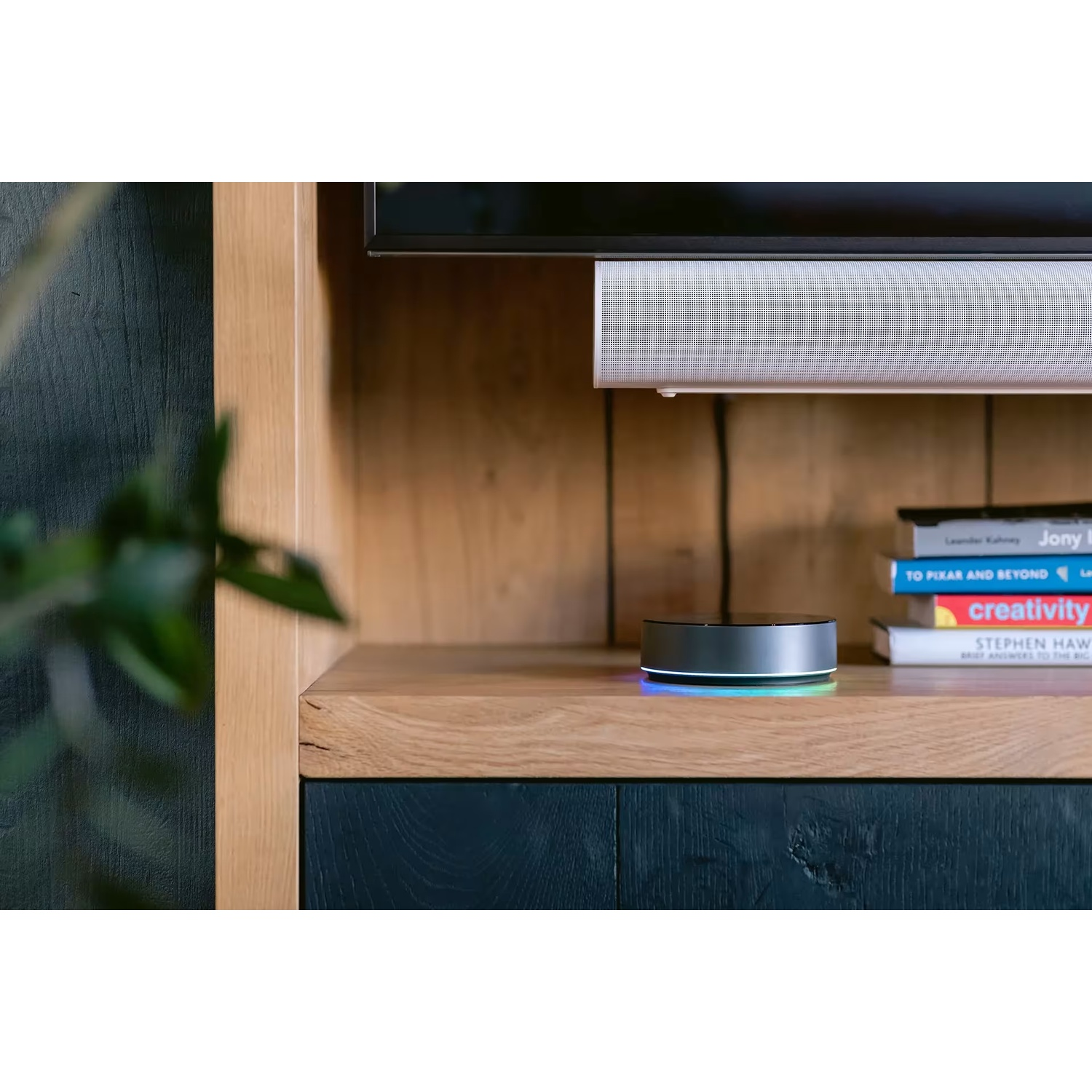 A sleek, silver smart speaker with a glowing blue and green light sits on a wooden shelf.