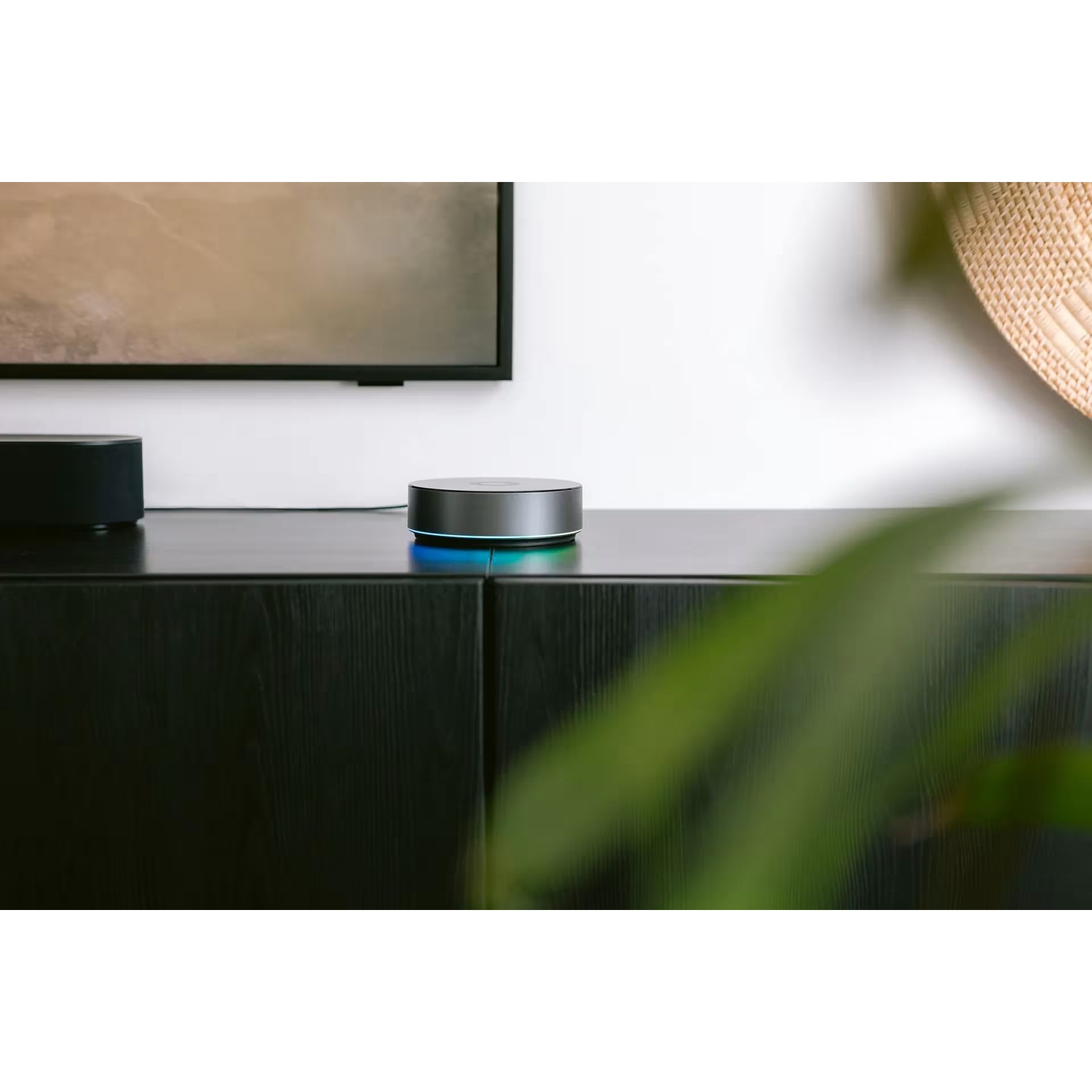 A silver smart speaker with blue light sits on a black cabinet, near a TV.