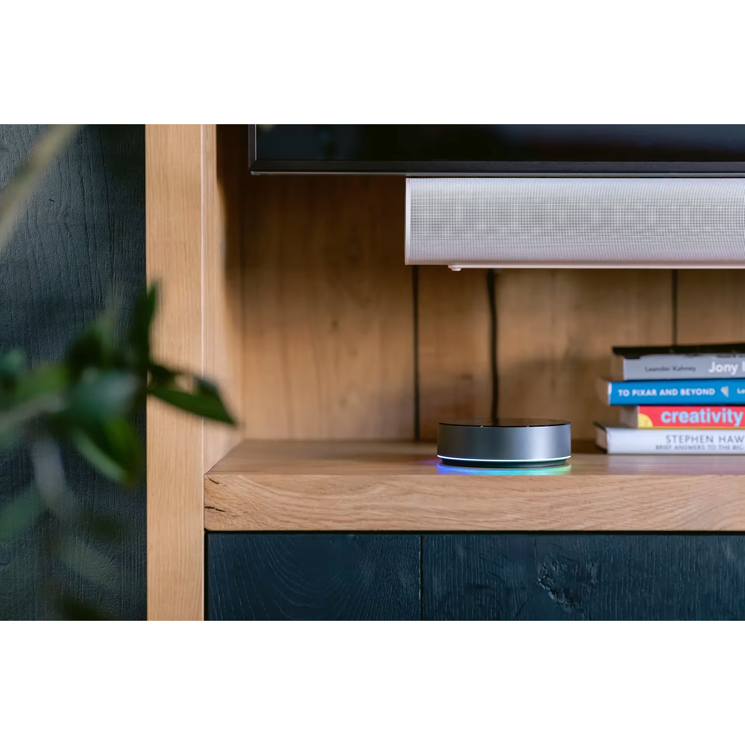 A round, silver smart speaker with a colored light ring sits on a wooden shelf.
