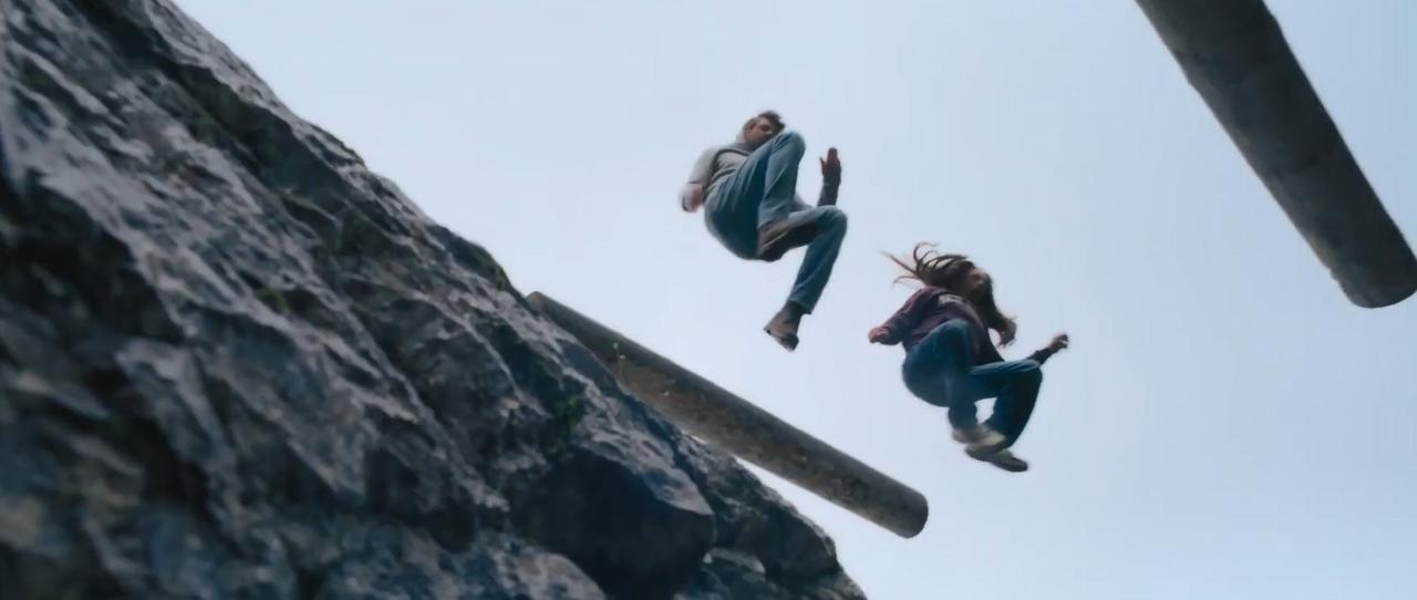 Zwei Personen springen von einer Klippe, umgeben von Felsen und Holzstämmen.