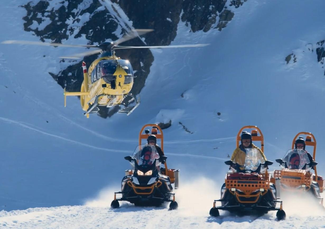 Ein gelber Hubschrauber fliegt über Schneemobile in einem verschneiten Berggebiet.