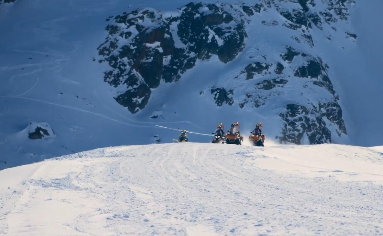 Schneemobile fahren auf einem verschneiten Berghang mit felsigen Klippen im Hintergrund.