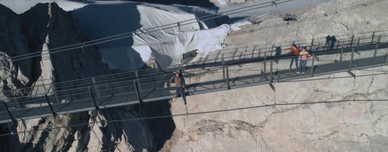 Personen gehen über eine Hängebrücke über eine felsige Gebirgsschlucht, weiße Planen sichtbar.
