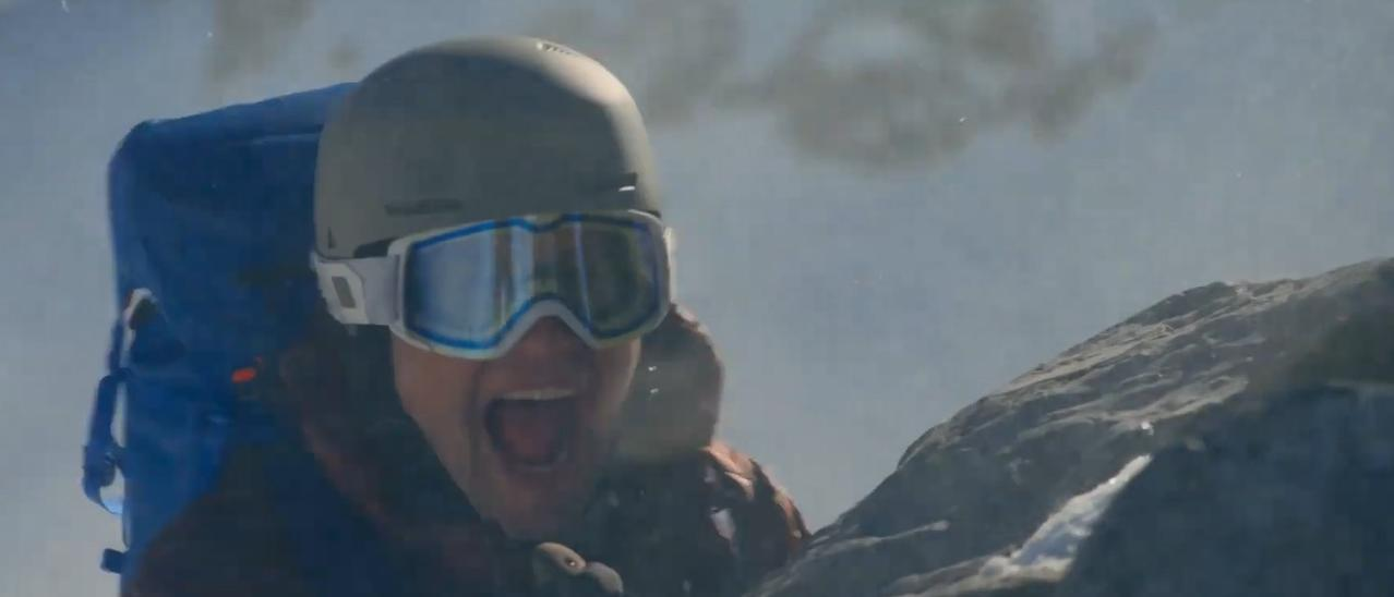 Eine Person mit Helm und Brille schreit beim Erklimmen eines felsigen, verschneiten Berges.