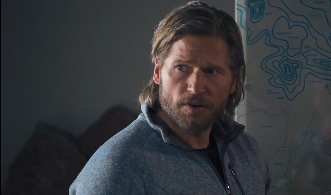 Ein Mann mit langen blonden Haaren und Bart trägt einen grauen Pullover neben einer Karte.