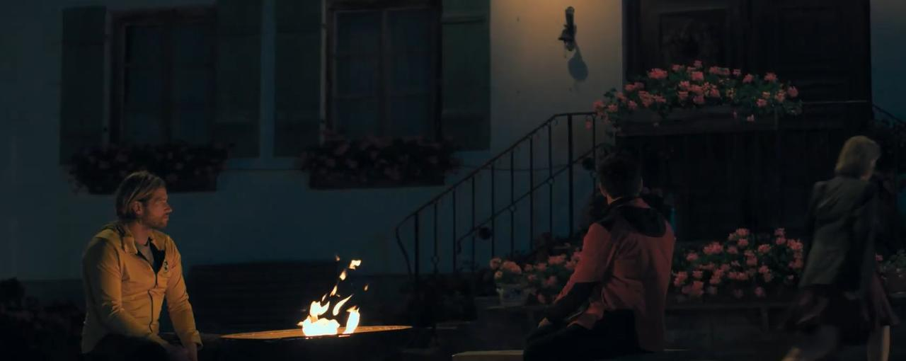 Drei Personen versammeln sich nachts um eine Feuerstelle vor einem Haus. Blumen schmücken den Eingang.