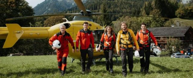 Vier Uniformierte gehen in einer Grasfläche mit Bergen in der Nähe eines gelben Hubschraubers.