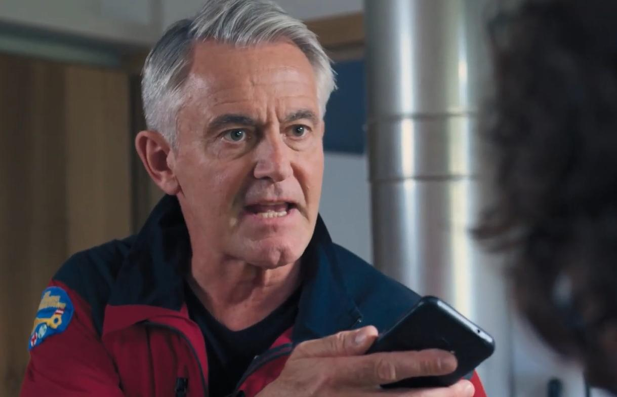 Ein älterer Mann mit grauen Haaren in einer rot-blauen Uniform hält ein Handy.