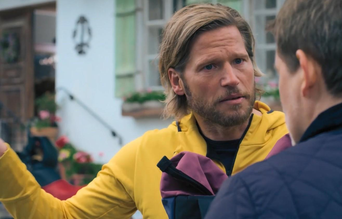 Ein Mann mit blondem Haar in einem gelben Hoodie spricht zu einer Person, die ihm den Rücken zuwendet.