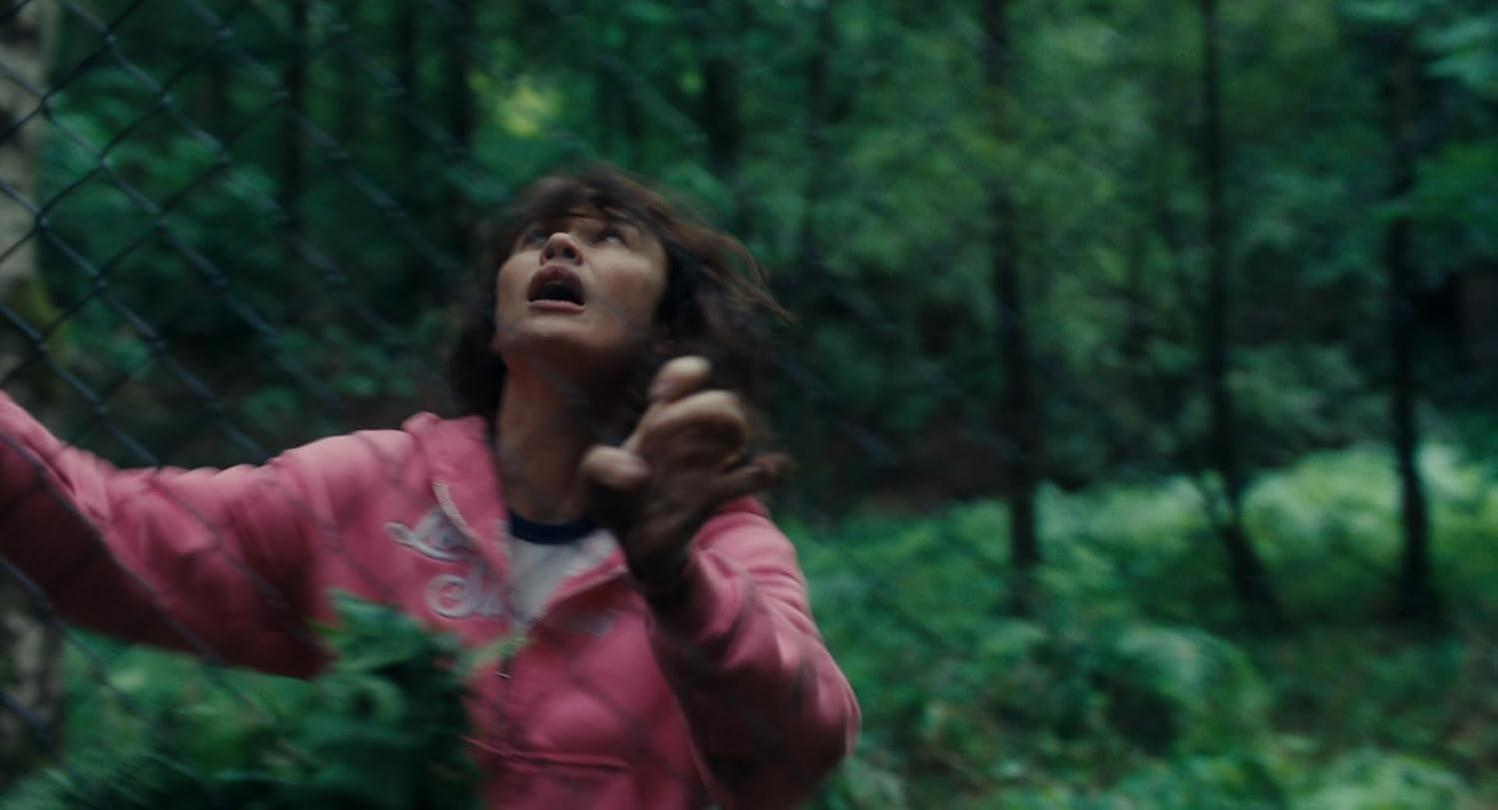 Eine Frau in einem rosa Hoodie blickt mit weit aufgerissenen Augen nach oben, umgeben von Bäumen.