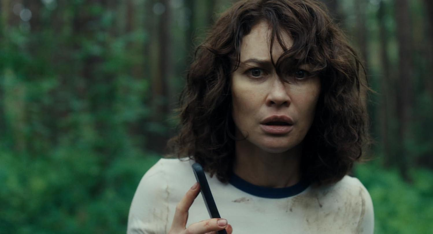 Eine Frau mit braunen Haaren hält ein Handy im Wald. Sie trägt ein weißes T-Shirt.