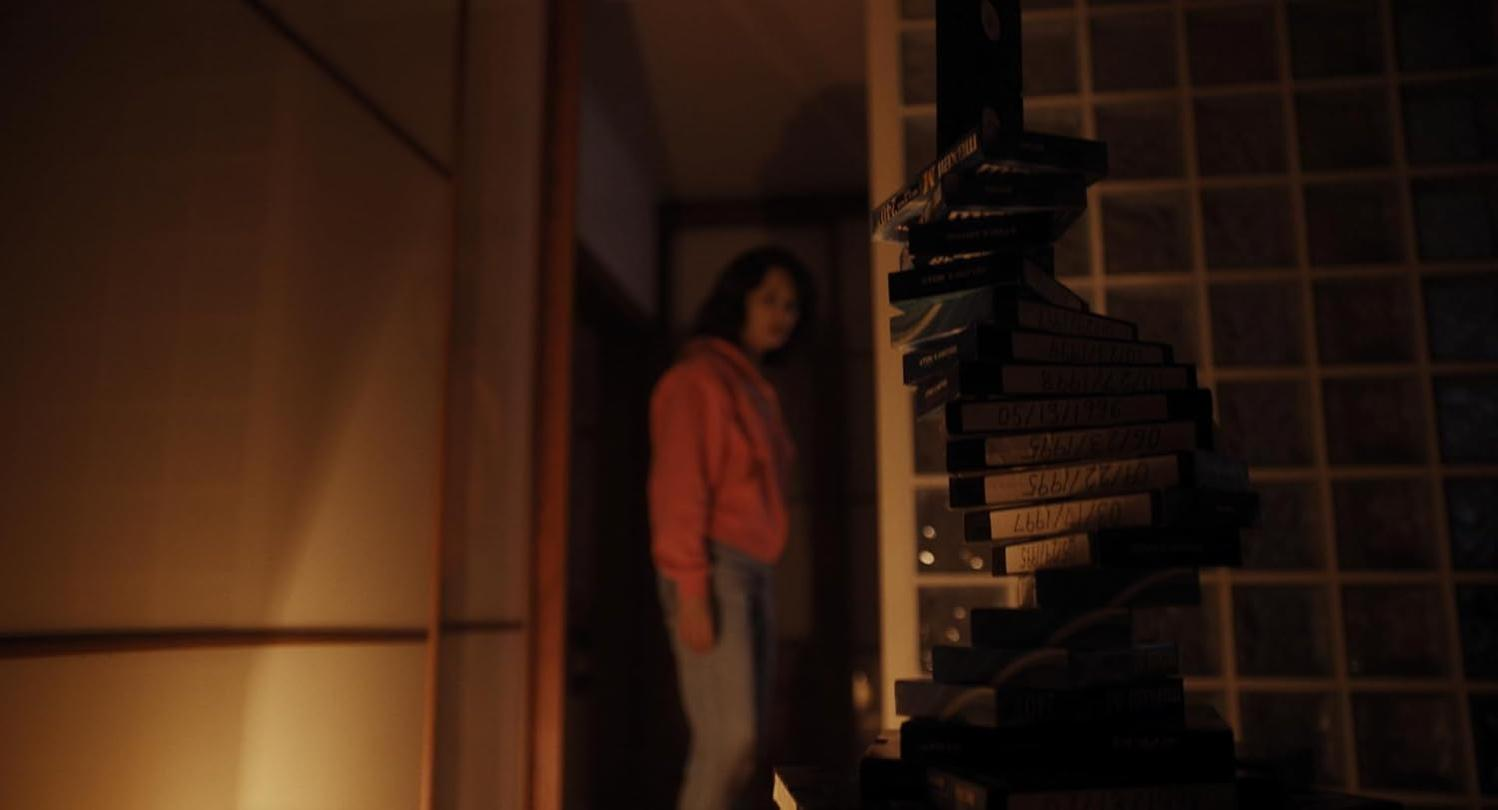 Eine Frau in einem roten Kapuzenpullover steht in einem schwach beleuchteten Raum neben einem hohen Stapel VHS-Kassetten.