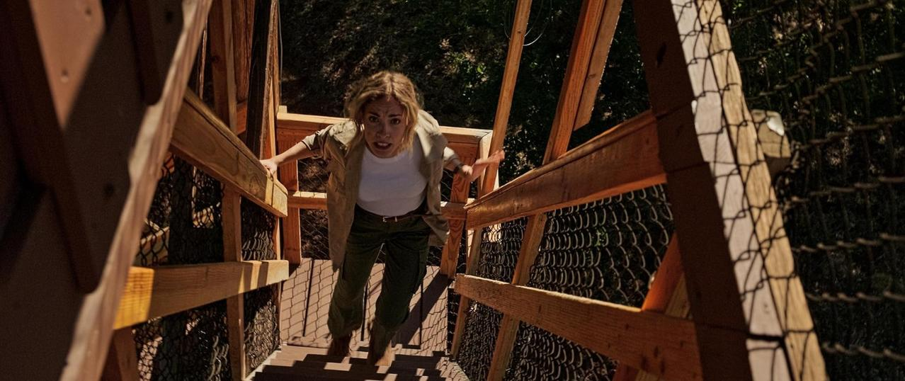 Eine Frau steigt eine Holztreppe hinauf, greift das Geländer und blickt ängstlich nach oben.
