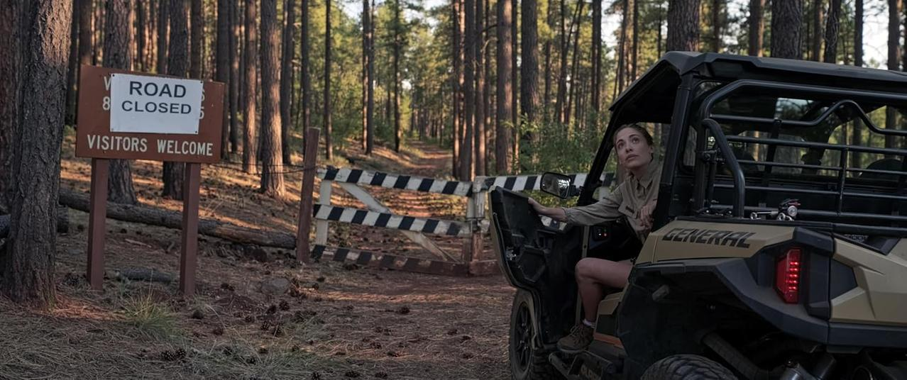 Eine Frau in einer sandfarbenen Uniform sitzt in einem sandfarbenen ATV neben einem "Road Closed" Schild im Wald.