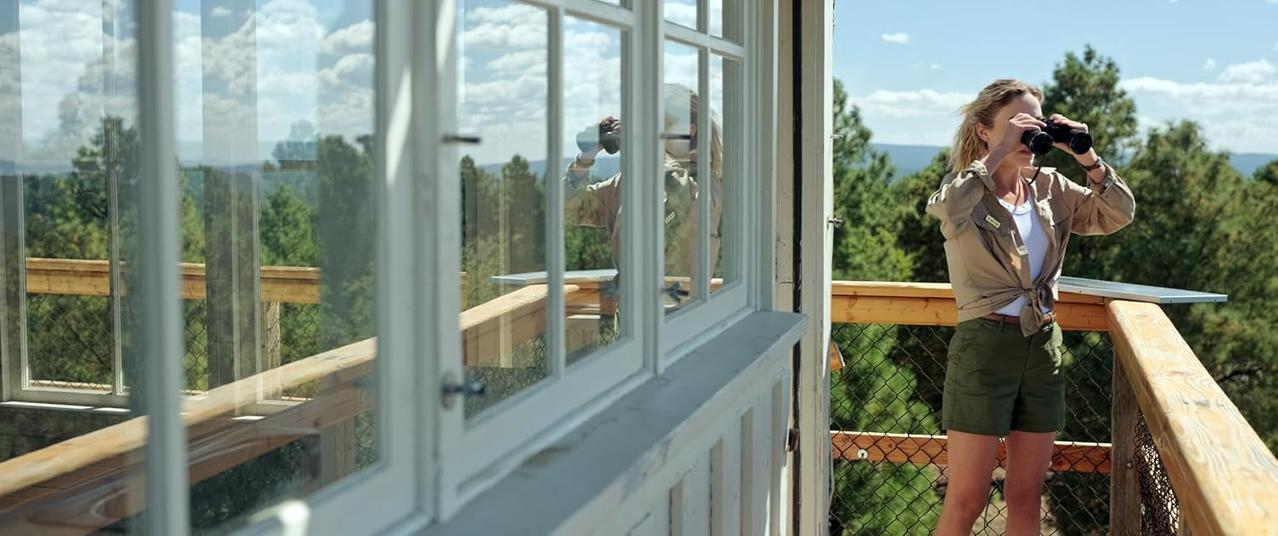 Frau mit Fernglas auf Holzbal kon mit Blick auf Wald.