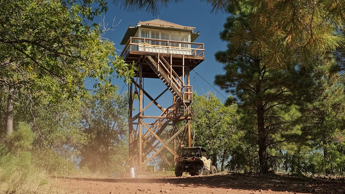 Hölzerner Brandwachturm mit Fahrzeug darunter, umgeben von Bäumen und staubiger Straße.