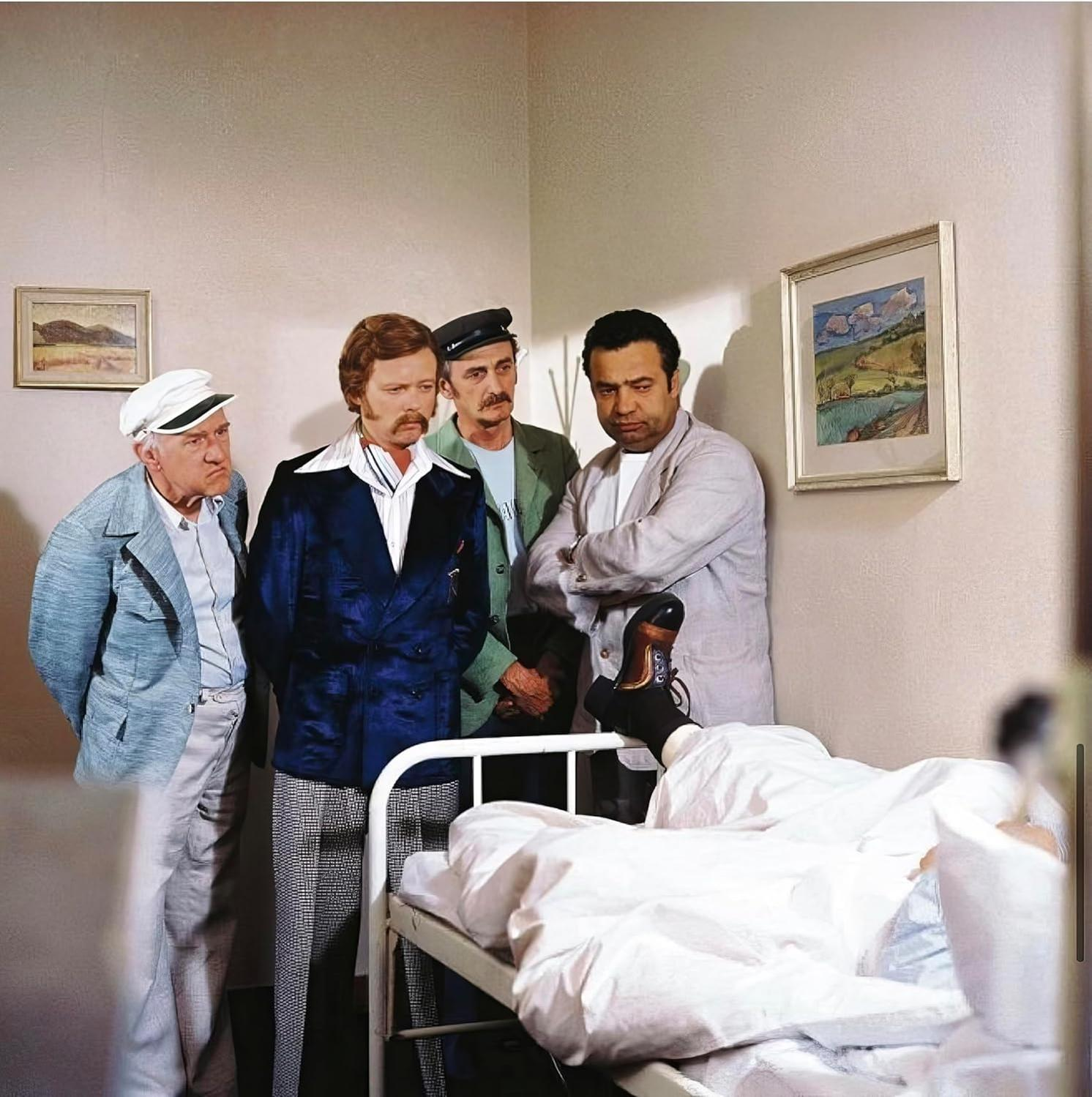 Vier Männer stehen in einem Raum mit einem Patienten im Bett. Zwei gerahmte Bilder hängen an der Wand.