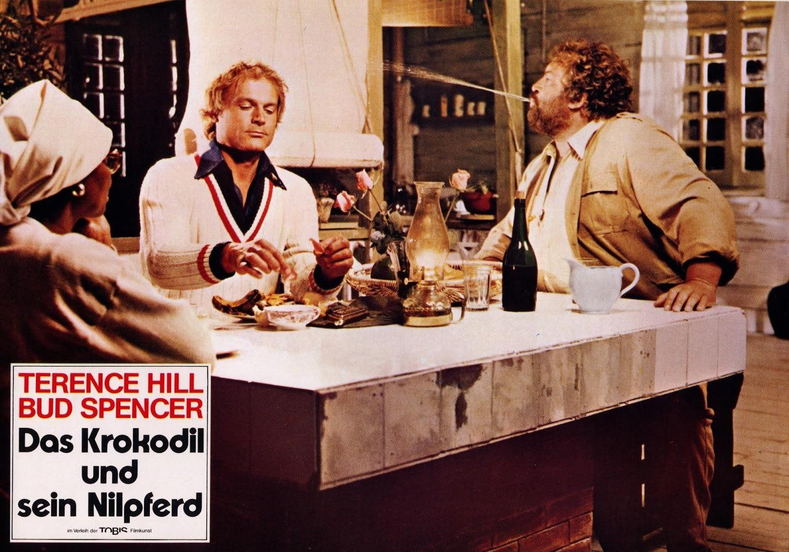 Drei Personen sitzen an einem Tisch, eine spuckt Wasser. Ein Filmplakat ist links.