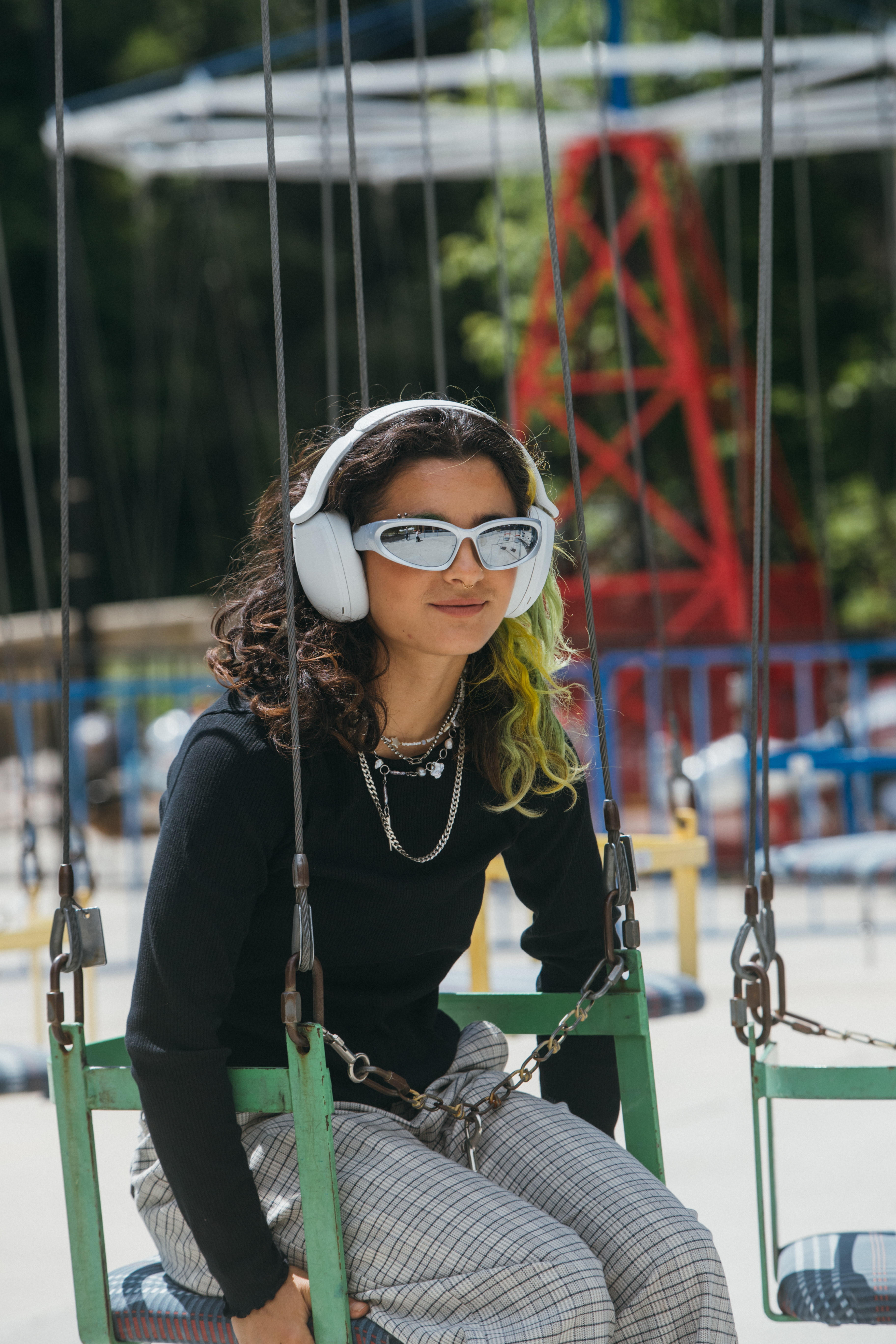 Eine junge Frau mit Kopfhörern und Sonnenbrille sitzt auf einer grünen Schaukel im Vergnügungspark.