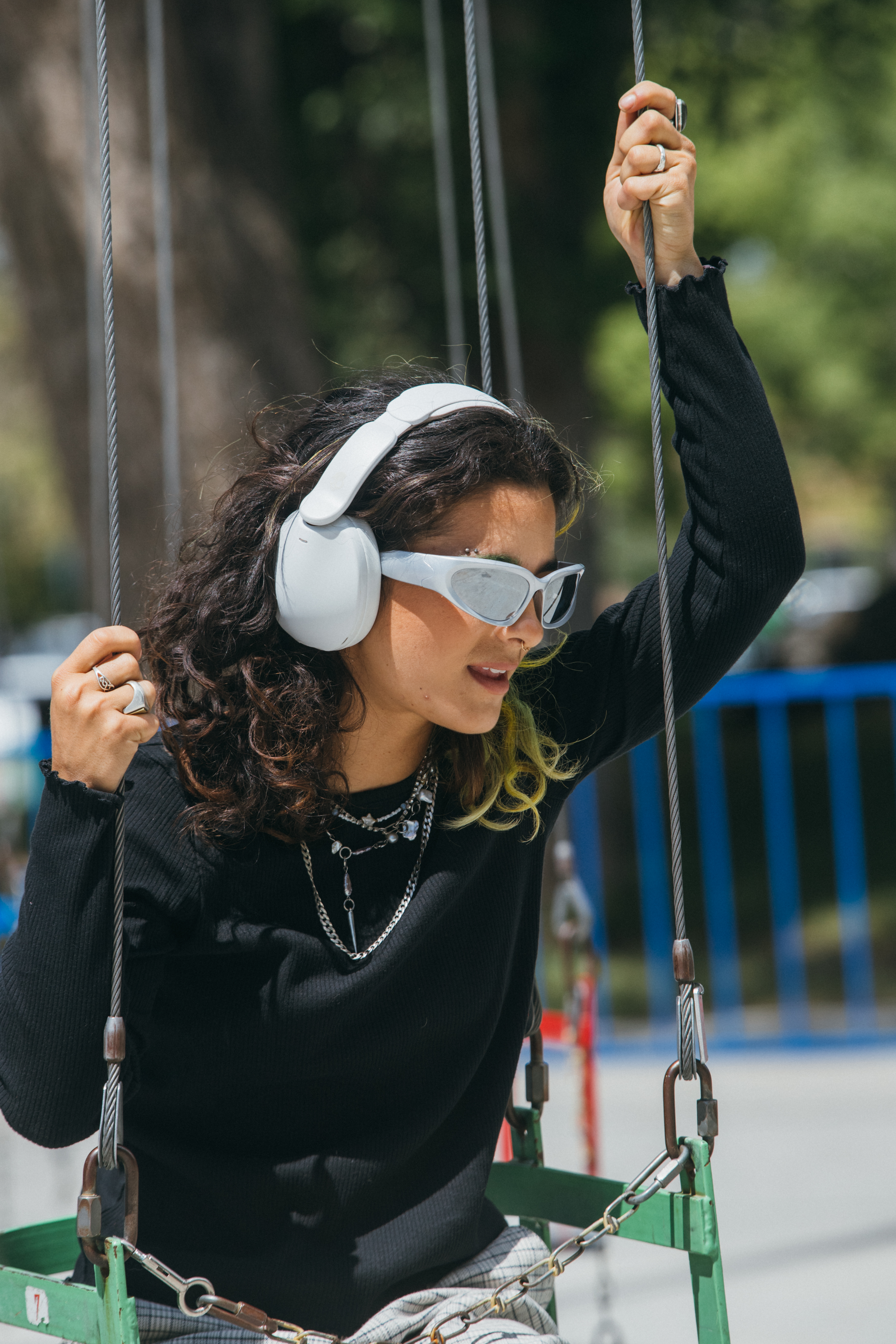 Junge Frau mit Kopfhörern und Sonnenbrille auf einer Schaukel, die sich an einer Kette festhält.