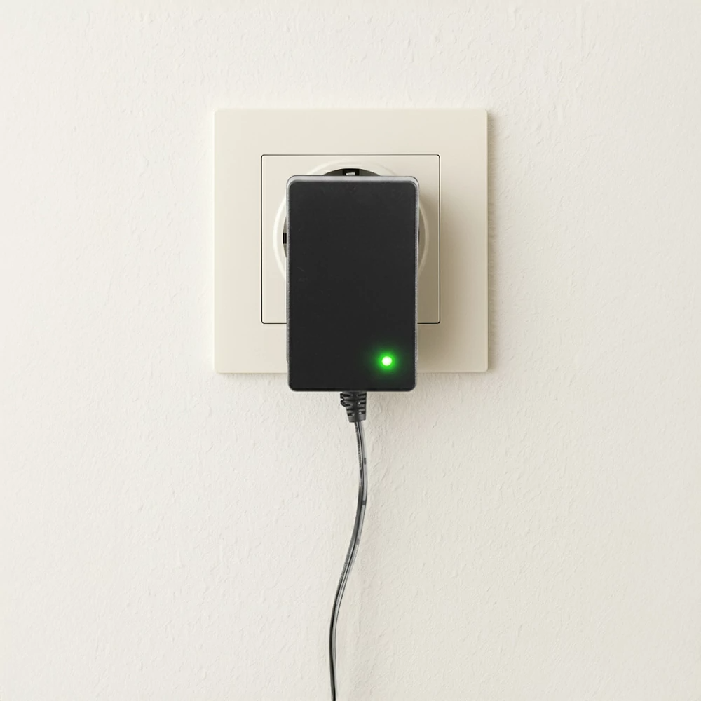 Fekete tápadapter egy fali aljzatban, zöld LED világít.