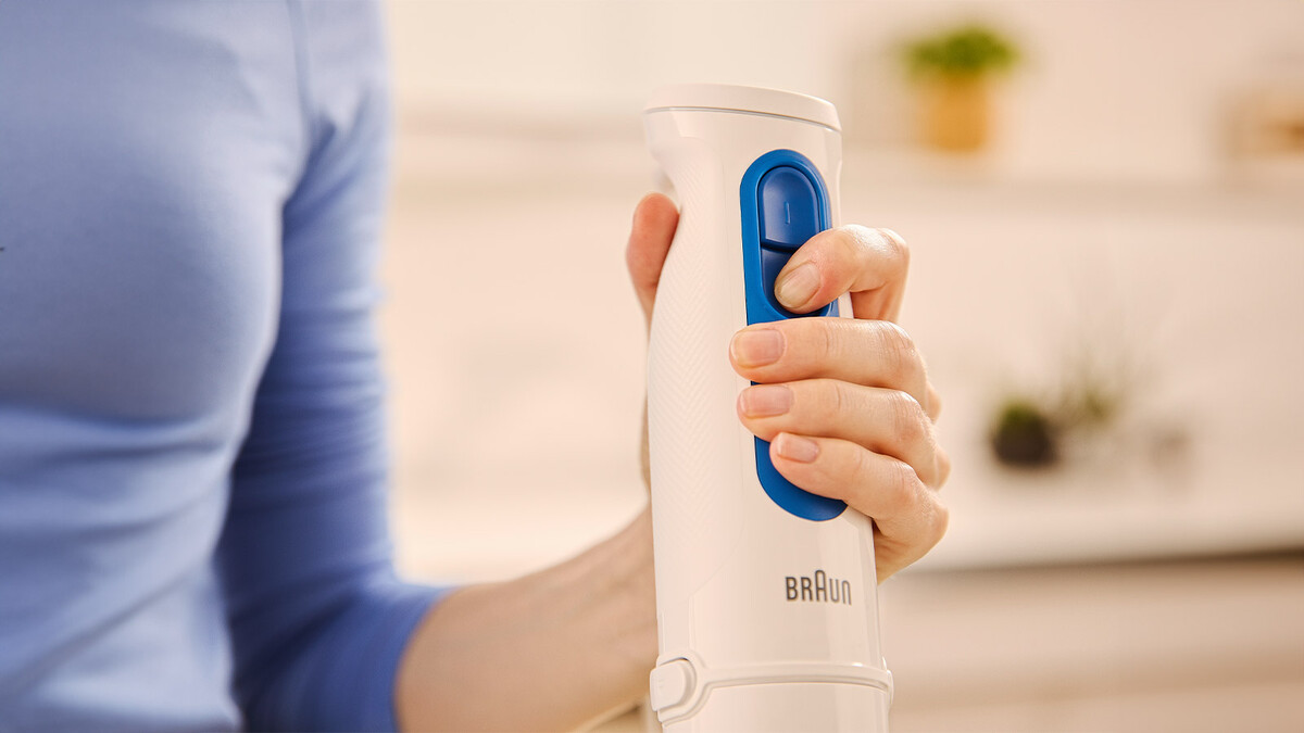 A person in a blue shirt holds a white Braun immersion blender with a blue button.