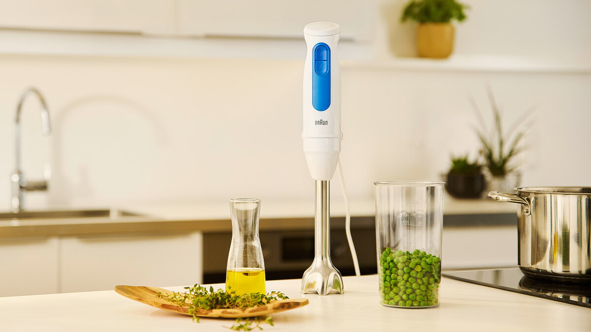 A hand blender stands beside oil, herbs, peas, and a pot in a kitchen.