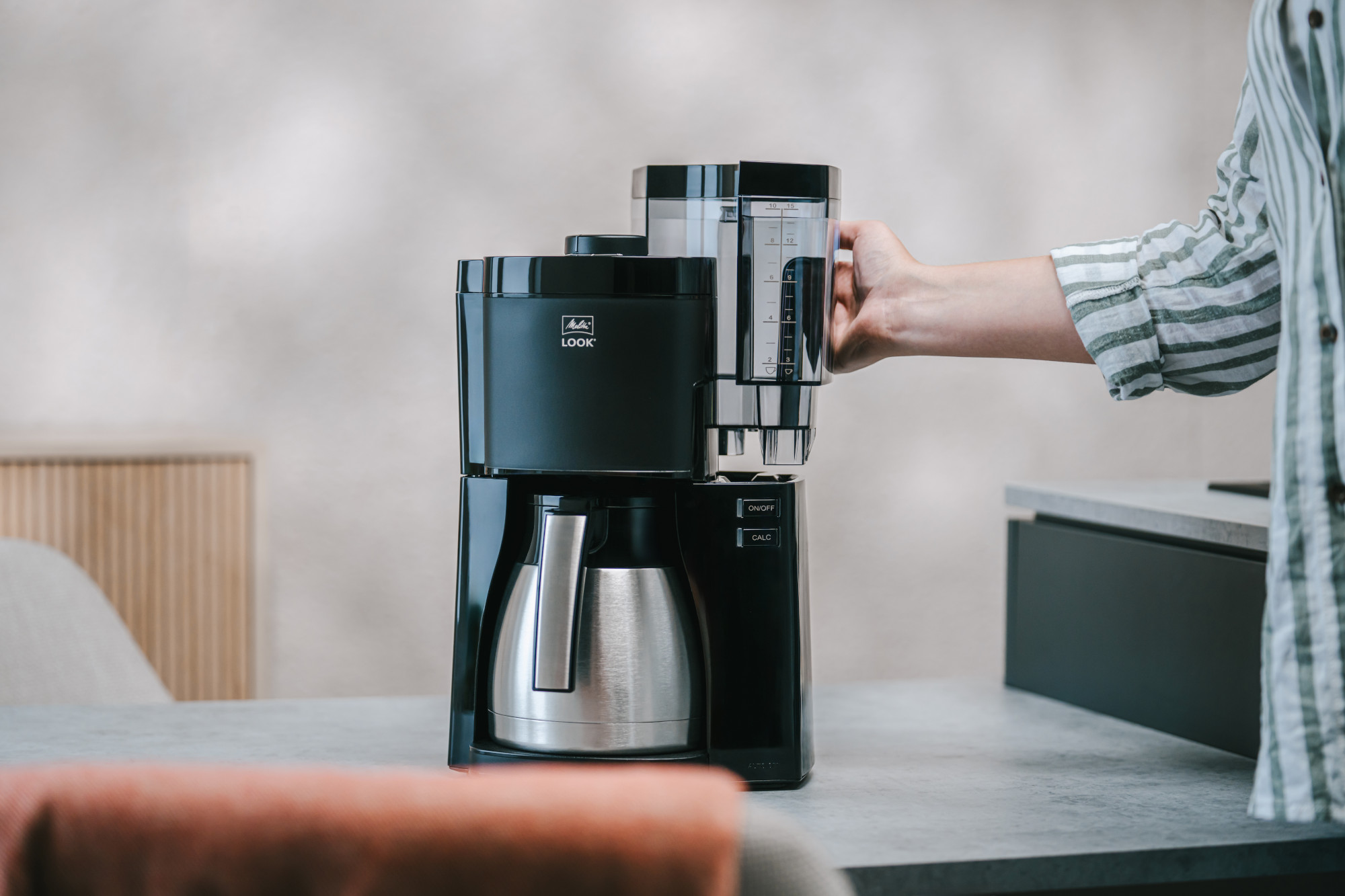Une cafetière Melitta noire avec une verseuse argentée sur une table. Une main remplit d'eau.