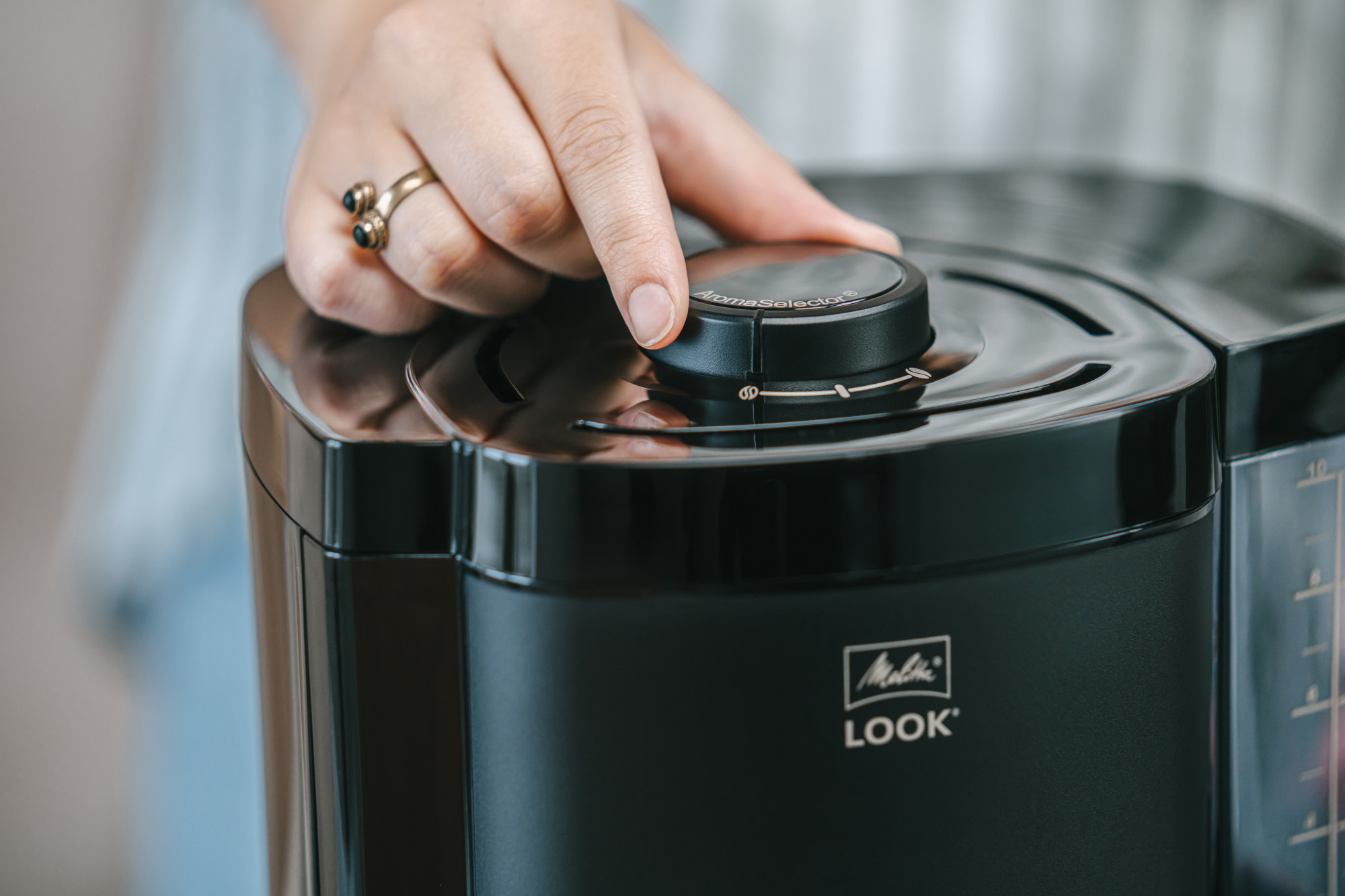 Une main avec une bague ajuste le sélecteur d'arôme sur une cafetière Melitta noire.