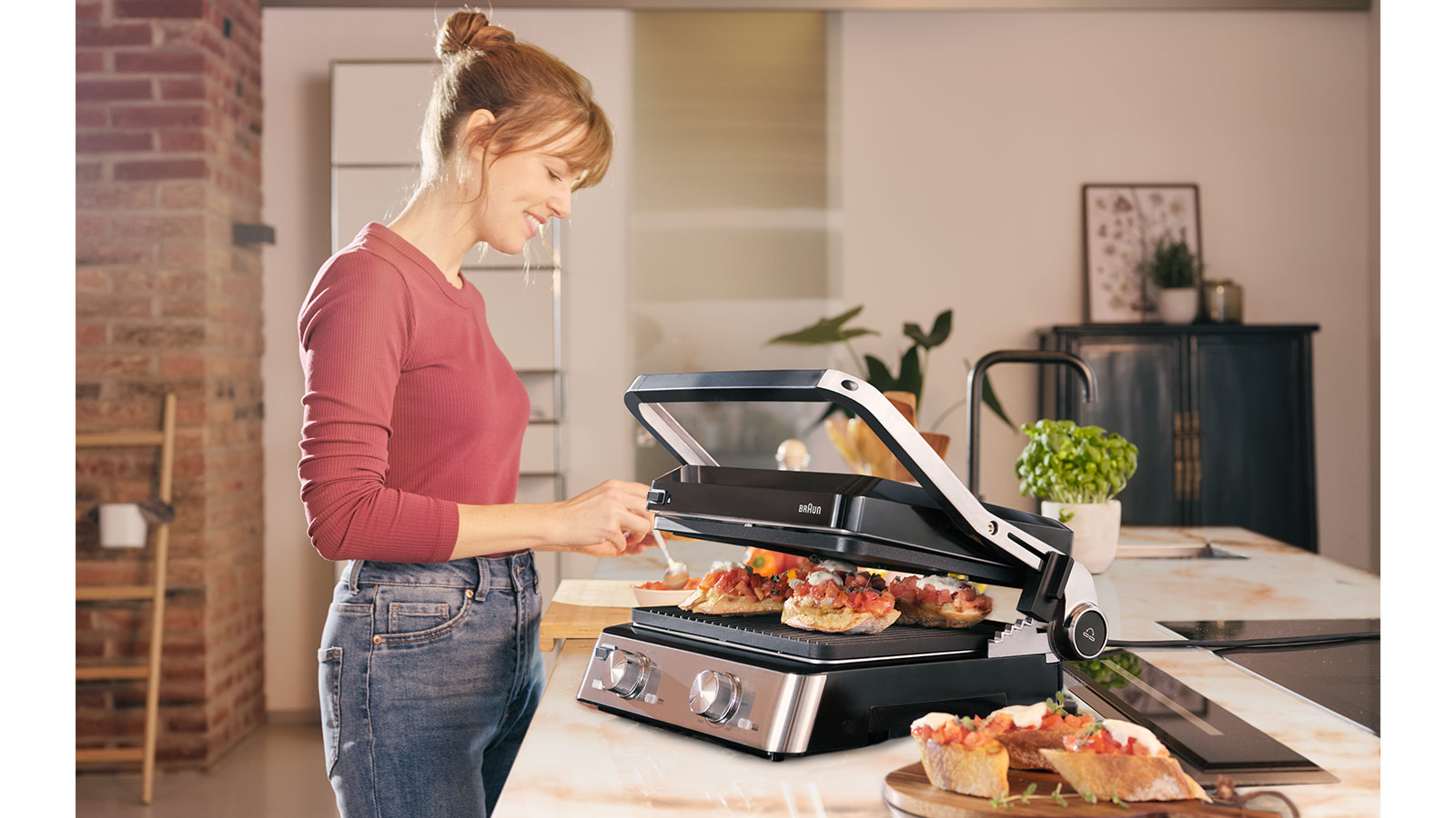 Frau in Jeans und pinkfarbenem Oberteil bereitet Essen auf einem Braun Grill in moderner Küche zu.