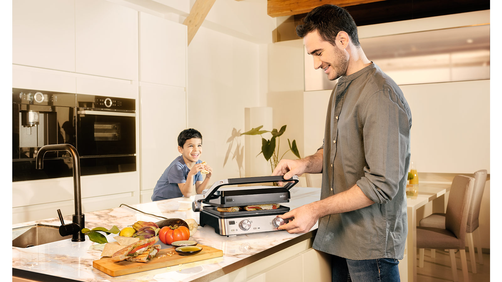 Mann und Junge in der Küche. Mann benutzt Grill. Junge isst Sandwich. Essen auf der Theke.