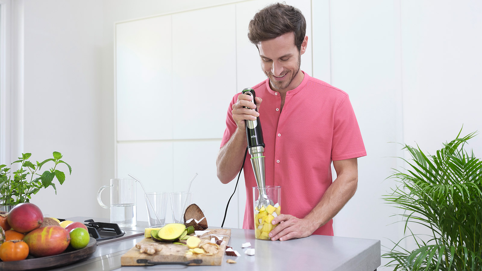 Mann in rosa Hemd mixt Obst mit Stabmixer in einem Glas. Obst auf der Theke.