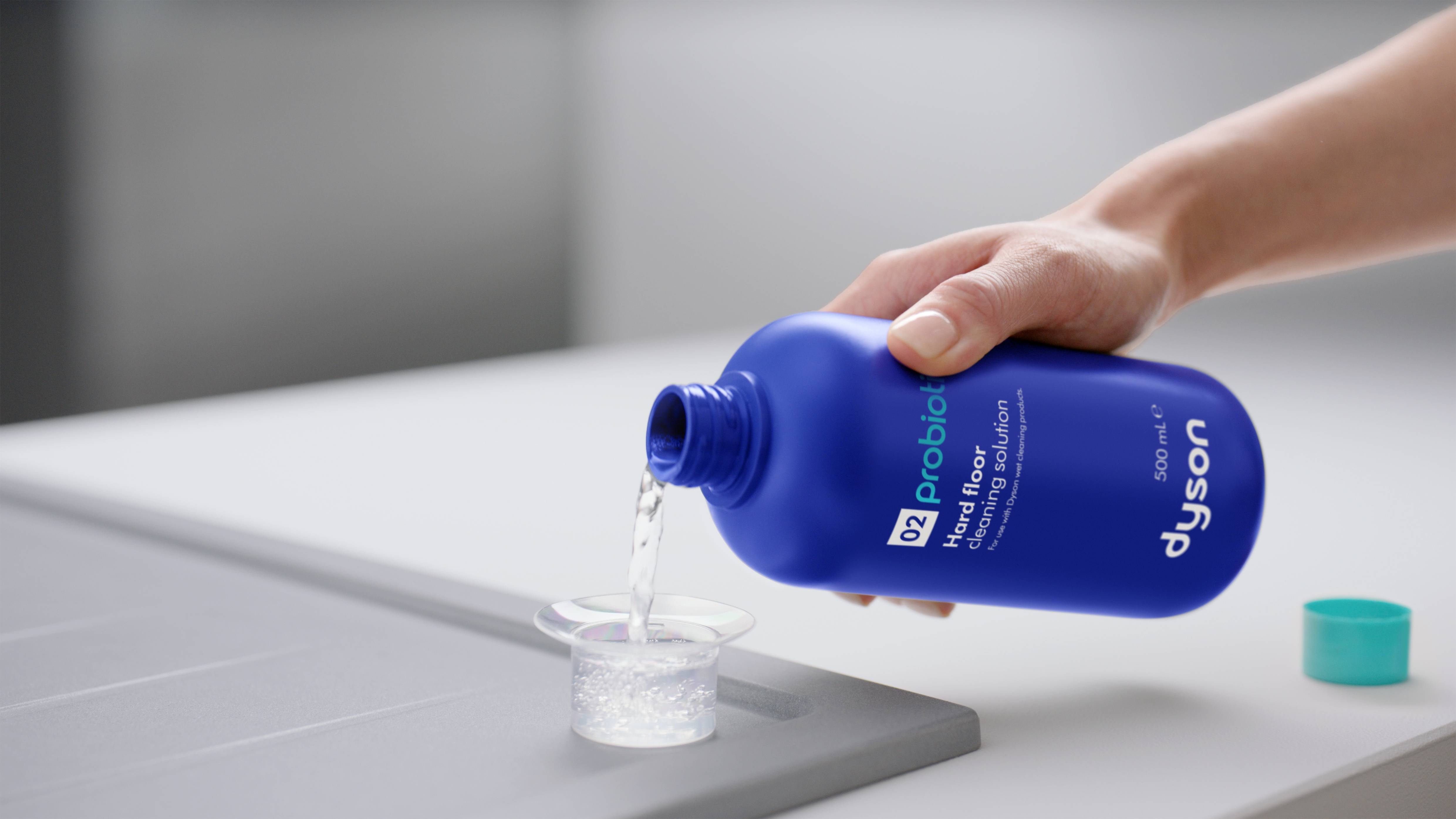 Una mano vierte limpiador de suelos Dyson azul en una tapa pequeña. Hay una tapa turquesa cerca.